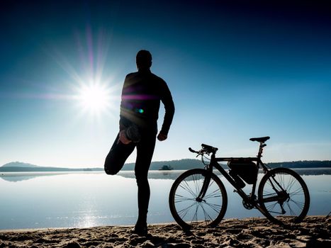 Outdoor silhouette of fit cyclist in cap at sunset with blurred reflection in water with ripples ride along seashore summer beach at evening horizon 