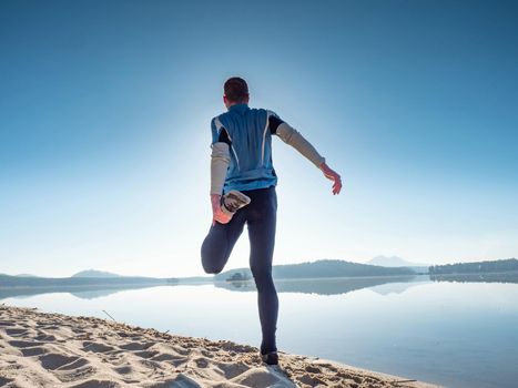 Athlete stretches his thigh muscles on the shore of the beach before running. Sunny morning training