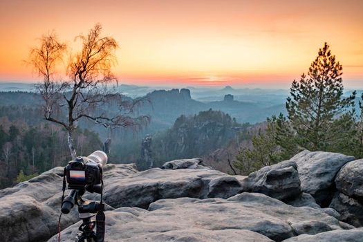 Camera on tripod is taking the sun sets just behind the Schrammsteine rock massif, Saxon Switzerland, Germany. View from the Carolafelsen viewpoint.