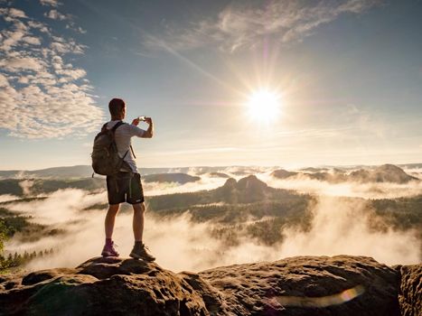 Man on top of a mountain watching the colorful sunset. Traveller with backpack photographing the beautiful nature scenics with smartphone while on vacation