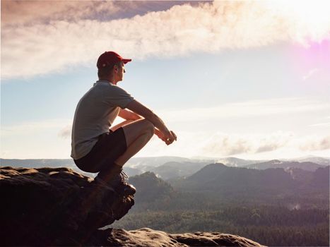 Man is squatting on the top of the mountain. Panoramic view of clouds below, sun in the sky and mountains. Active recreation, climbing, trekking and hiking 