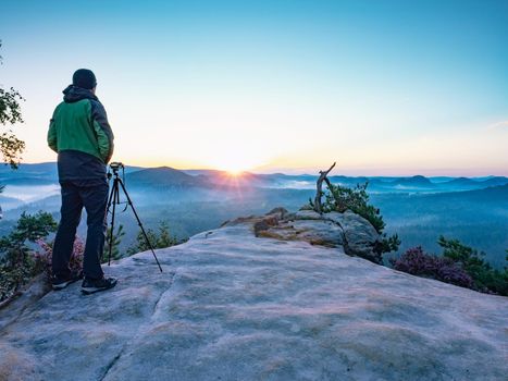 Artist with camera and tripod watch sunrise on rocky summit. The photographer enjoy misty autumn sunrise on sharp cliff in  Saxon Switzerland National Park.