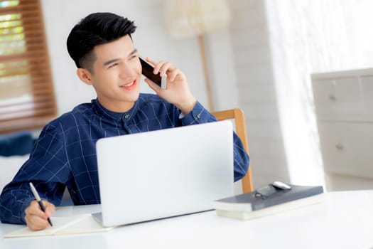 Young asian man talking phone and work from home with laptop computer, freelance male writing on notebook and speak on smartphone, stay home, domestic life, business and communication concept.
