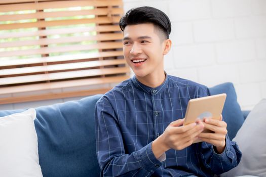Young attractive asian man resting using browsing tablet computer on sofa at home, happy male sitting on couch relax reading digital gadget at house, communication and lifestyle concept.