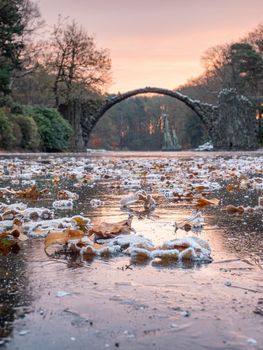 Frosty night with full moon light. Frozen lake with colorful leaves, round Rakotzbruecke, the Devil's bridge,  in light morning mist.  Kromlau park, Germany