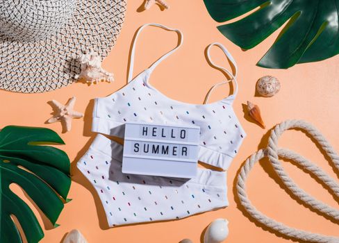 Summer and vacation concept. Words Hello Summer on the lightbox with swimming suit, tropical leaves and seashells flat lay on orange background