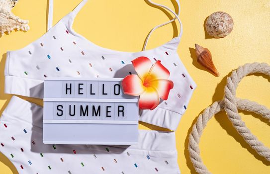 Summer and vacation concept. Words Hello Summer on the lightbox with swimming suit, tropical leaves and seashells flat lay on orange background