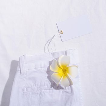 Summer and vacation concept. White summer jeans with plumeria flower on white background with blank price tag. Mock up design