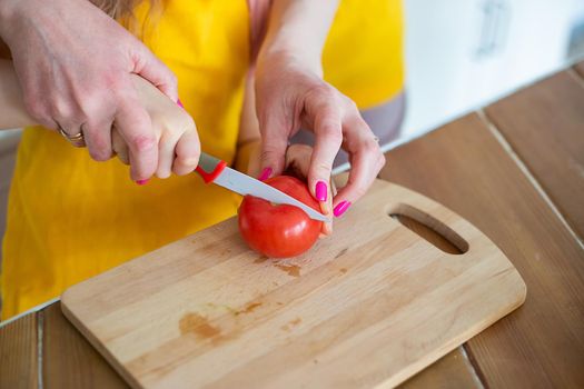 Cropped image of Young Mother Helping Daughter to cut tomato with kitchen knife on cutting board, teaching child to cook