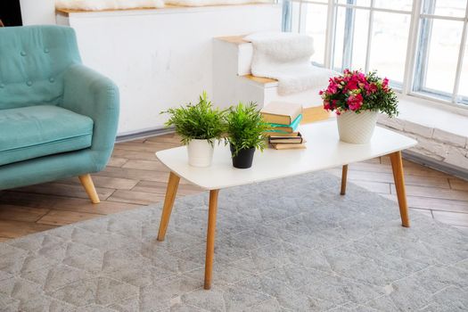 A white coffee table stands near a window with potted plants and books on it in a bright Scandinavian-style living room