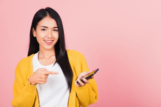 Portrait Asian happy beautiful young woman smile making finger pointing on smartphone in hand shopping online application looking to camera isolated, studio shot pink background with copy space