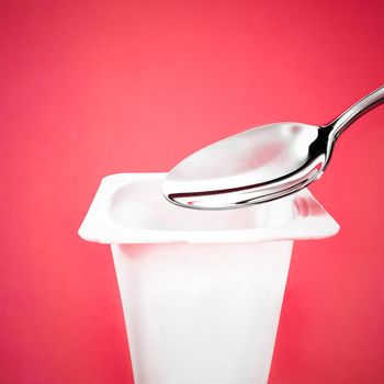 Yogurt cup and silver spoon on red background, white plastic container with yoghurt cream, fresh dairy product for healthy diet and nutrition balance.