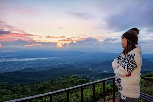 Woman tourist in a sweater dress standing watching the beautiful nature landscape of the forest and mountain during the sunrise on the high peak in the morning at Phu Thap Boek Viewpoint Phetchabun, Thailand