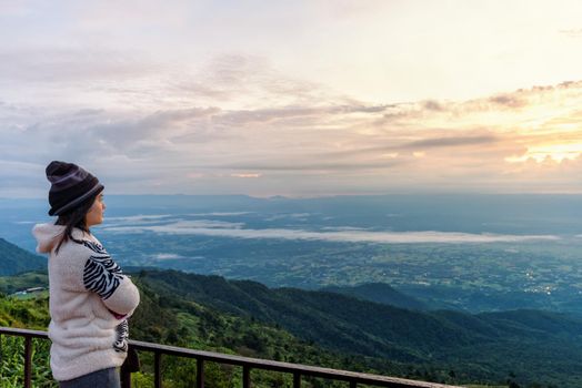 Woman tourist in a sweater dress standing watching the beautiful nature landscape of the forest and mountain during the sunrise on the high peak in the morning at Phu Thap Boek Viewpoint Phetchabun, Thailand