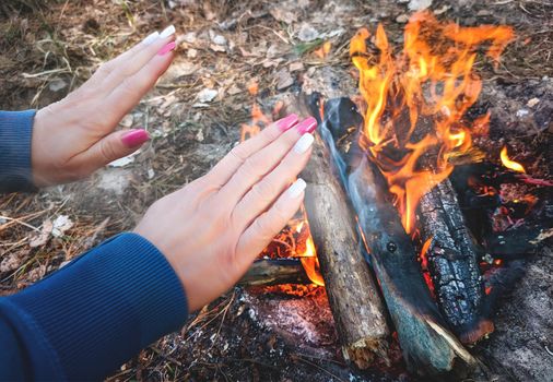 woman is making fire while camping outdoors, at the forest - warming up her hands near the fire