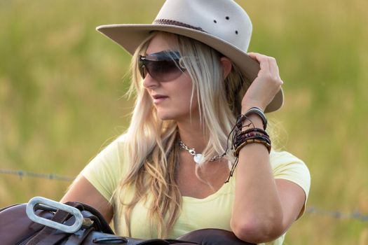 Casual dressed woman with akubra hat, oversized sunglasses leaning by her saddle.  She is wearing leather and braided bangles