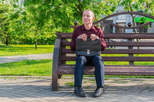 An office worker in a business suit with a suitcase and documents sits with a stupid, confused view on a bench in a city park and expects a meeting