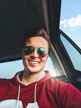 A young smiled man is sitting in the back seat of a taxi cab going to the airport and taking selfie