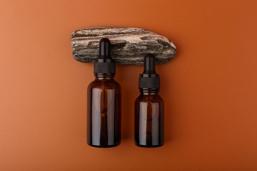 Two dark transparent glass bottles with skin serum or oil for manicure on natural stone against brown background .