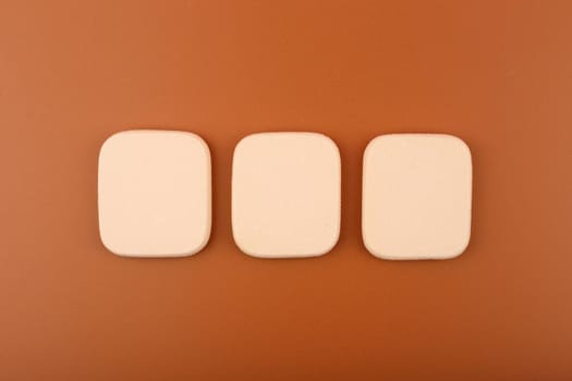 Three beige square shaped make up sponges on brown background. Concept of make up and visage for dark skin tones