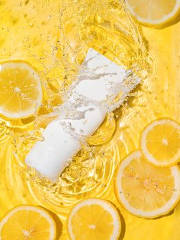 Cosmetic empty white tube in clean transparent water with lemon slices over yellow background. Water splashing on surface, sunlight. Vertical flatlay,copy space. Beauty mockup for cream or moisturizer