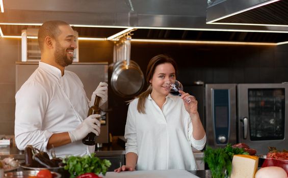 Handsome young African chef is cooking together with Caucasian girlfriend in the kitchen using red wine ingredient. A cook teaches a girl how to cook. Man and woman cooking in professional kitchen.