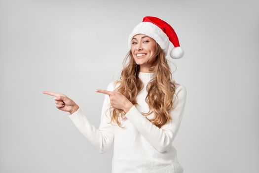 Woman christmas Santa Hat sweater white studio background Beautiful caucasian female curly hair portrait Happy person positive emotion Holiday concept Shows finger on side cope space for advertise