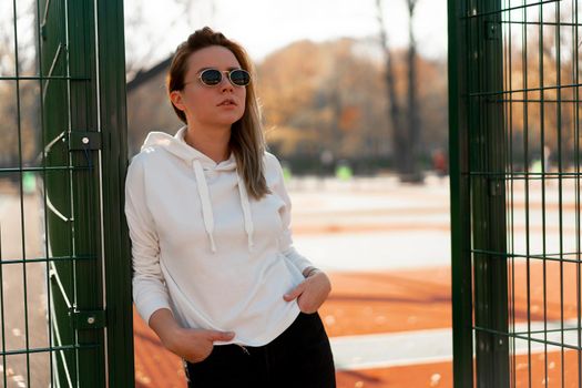 Outdoor close up portrait of young beautiful woman with long hair in sunglasses, dressed in a white hoodie sweater, near the sportsground. youth culture summer pastime