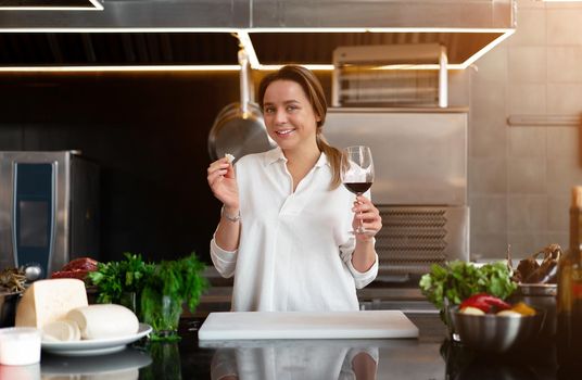 Beautiful young caucasian girl standing in kitchen in a white uniform smiling and tasting red wine Cute woman 30s years old in white shirt with food Ingredients cheese meat vegetables drinking