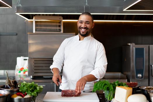 Handsome young African chef standing in professional kitchen in restaurant preparing a meal of meat and cheese vegetables. Portrait of man in cook uniform Cuts meat with a metal knife.