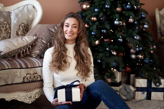 Christmas. Woman dressed white sweater and jeans sitting on the floor near christmas tree with present box Caucasian female middle age opens gifts winter holiday at home interior New year mood