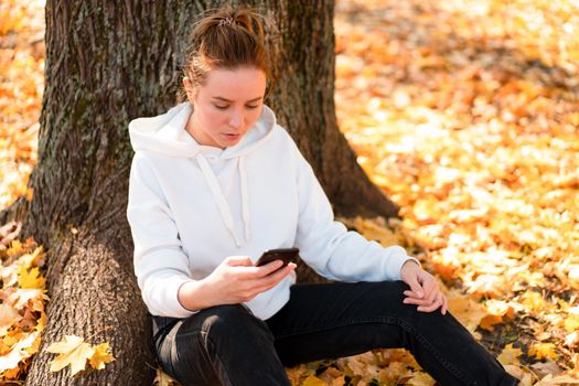 Woman in a white hoodie sweater with a hood sits on ground in the park and holding a cellphone in her hands. cute girl use smartphone for communication Female texting a message Communicaion concept