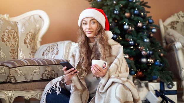 Christmas Woman dressed white sweater Santa hat sitting on floor near christmas tree wrapped herself blanket Caucasian female relaxing winter holiday plaid drink hot coffee using smartphone