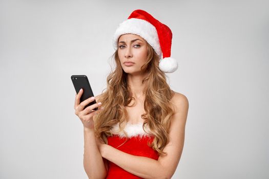 Woman christmas Santa Hat white studio background with smartphone in hand Beautiful caucasian female curly hair portrait Serious person sad emotion Holiday concept with copy space bad mood melancholy