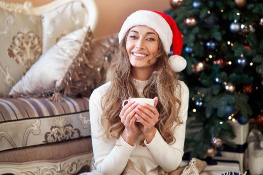 Christmas Woman dressed white sweater Santa hat sitting on floor near christmas tree wrapped herself blanket Caucasian female relaxing winter holiday at home interior with plaid drink hot coffee