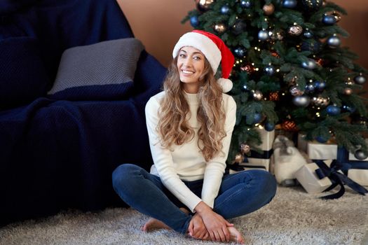 Christmas. Woman dressed white sweater jeans santa hat sitting on the floor near christmas tree with present box Caucasian female middle age relaxing winter holiday at home interior New year mood