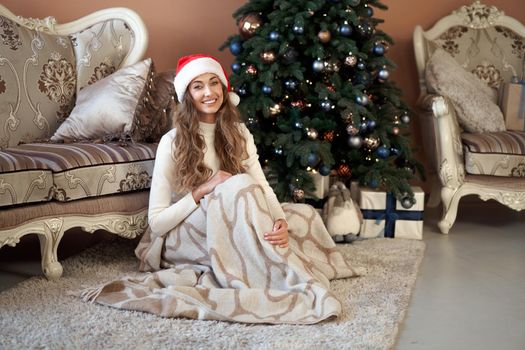 Christmas. Woaman dressed white sweater Santa hat sitting on the floor near christmas tree wrapped herself blanket. Caucasian female middle age relaxing winter holiday at home interior with plaid