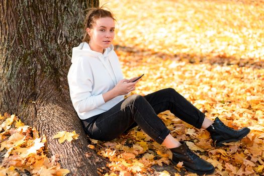 Woman in a white hoodie sweater with a hood sits on ground in the park and holding a cellphone in her hands. cute girl use smartphone for communication Female texting a message Communicaion concept