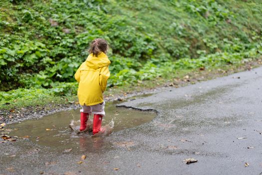 Playful girl wearing yellow raincoat while jumping in puddle during rainfall Happy childhood