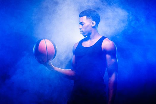 Basketball player holding a ball against blue fog background. African american man looking at basket.