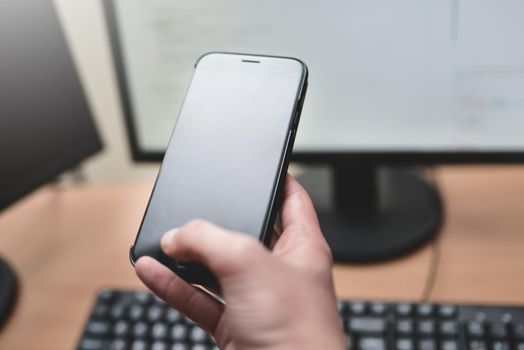 Person holding a smart phone and pointing her finger at the blank screen against a computer keyboard,monitor on wooden table. business and technology concept