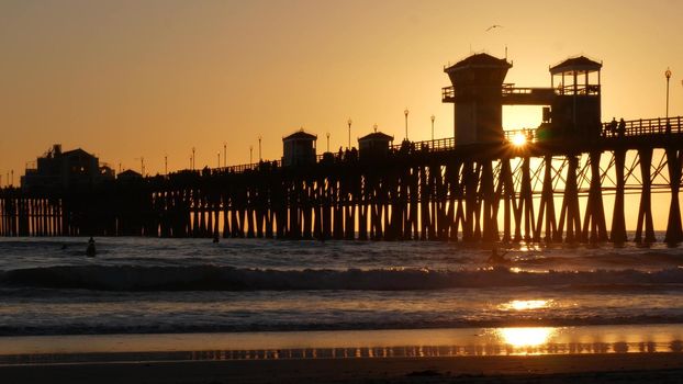 Wooden pier silhouette at sunset, California USA, Oceanside. Waterfront surfing resort, pacific ocean tropical beach. Summertime sea coastline vacations atmosphere. Surfers waiting for wave in water.