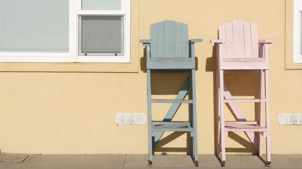 Two vintage wooden lifeguard chairs by yellow wall, California USA. Empty life guard retro high seat by ocean sea beach. Minimal simple trendy pop atmosphere, summertime aesthetic. Los Angeles vibes.