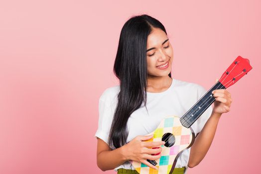 Portrait of happy Asian beautiful young woman teen confident smiling face hold acoustic Ukulele guitar, female playing Hawaiian small guitar, studio shot isolated on pink background, with copy space