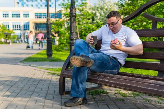 A young man in white sitting on a park bench accidentally spills hot coffee on his pants during lunch