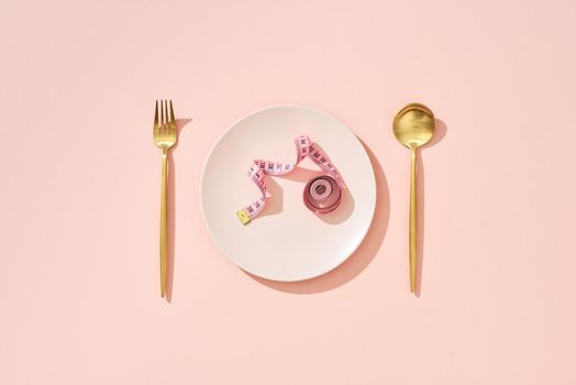Roll of measuring tape on a pink dinner plate, table knife and fork on a pink background.