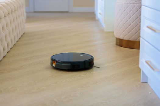 The circular robot vacuum cleaner collects debris from the living room floor. Black color.