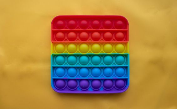 A rainbow toy lies against a background of yellow fabric. Blue and Orange Funny Interactive Anti-Stress Silicone Toys. Pop it.