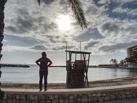 Woman backpacker walk on coastline promenade at lifeguard tower. Swimming bay at hotel beach, summer vacation resort, Mallorca