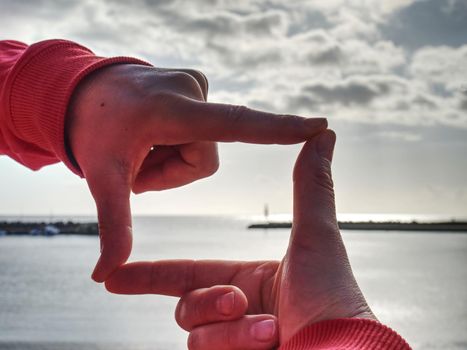 Fingers gesturing picture frame on sunrise seaside. Sunlight on beach. Sunset at the sea. Hand-made gesture in love concept, female  wrist